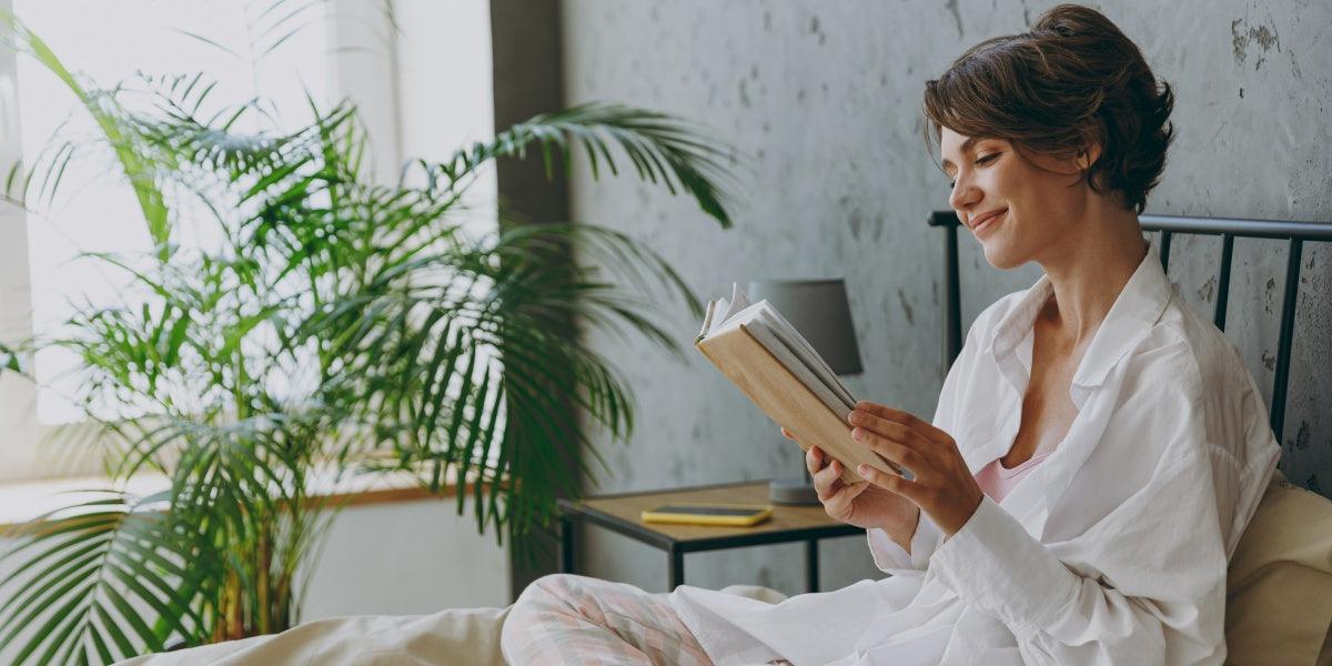 Side view young calm happy woman wear white shirt pajama she lying in bed read book study rest relax spend time in bedroom lounge home in own room hotel wake up dream be lost in reverie good mood day.