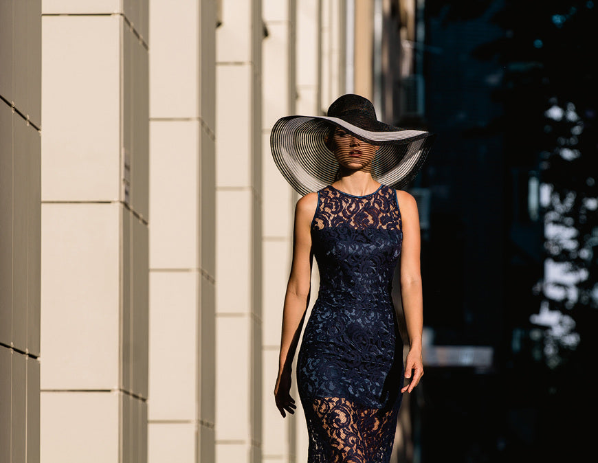Young-woman-in-black-lace-dress-and-a-hat-with-a-wide-brim.-She-walks-down-street-along-building.