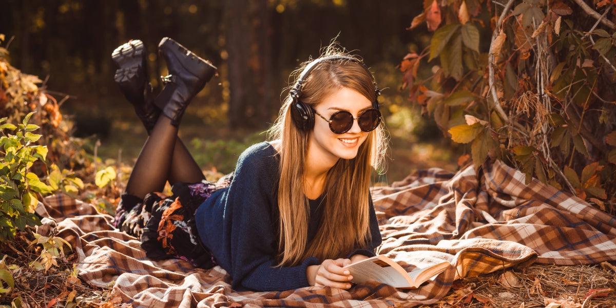 Young girl in headphones listening to music lying on a rug under the tree in the autumn forest. Girl in sunglasses dressed in a blue sweater and holding a book