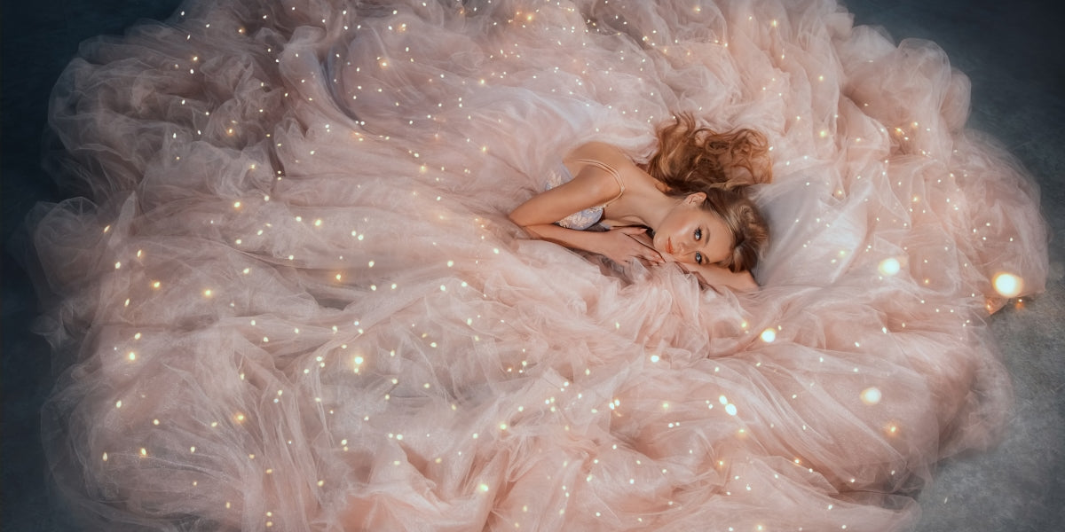 Fairy queen fashion model in luxurious shining pink dress posing in studio. Princess girl, peach outfit with sequins lies in fabrics of outfit, long skirt gown. Fantasy woman sleeping beauty woke up