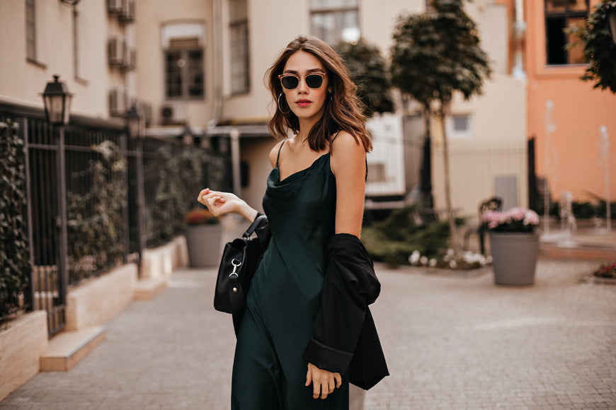 Stylish-pale-brunette-in-green-long-dress-black-coat-and-sunglasses-standing-on-the-street-during-daytime-with-bright-city-buildings-in-the-background