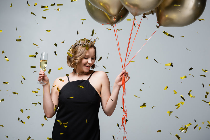 happy-plus-size-woman-in-slip-dress-and-crown-holding-balloons-and-glass-of-champagne-near-confetti-on-grey
